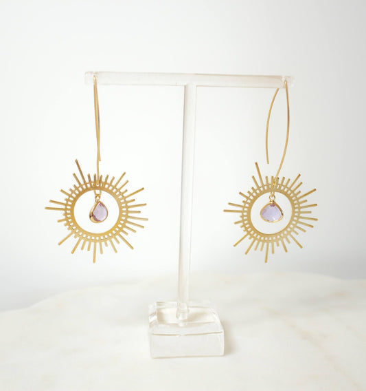 Gold earrings with sunburst design on a clear stand against a white background